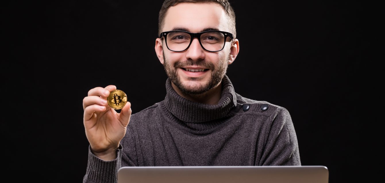 Portrait of a happy businessman showing bitcoin while sitting at desk with laptop computer isolated over black background