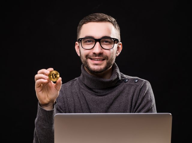 Portrait of a happy businessman showing bitcoin while sitting at desk with laptop computer isolated over black background